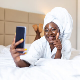 Smiling african woman lying on bed in bathrobe with mobile phone taking a selfie. African woman relaxing on the bed after bath and looking at the phone camera taking a selfie showing thumbs up